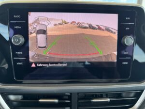Rückfahrkamera-Display zeigt Parkplatz, Auto mit Hilfslinien; Warnung: "Fahrweg kontrollieren!".