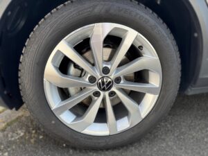 Volkswagen-Rad mit silbernen Speichen auf schwarzem Reifen vor grauem Hintergrund.
