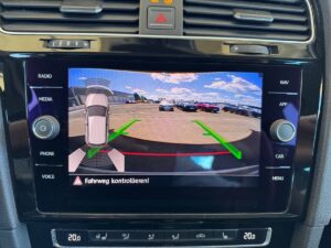 Autodisplay zeigt Rückfahrkameraansicht mit parkenden Autos und Anleitungspfeilen, sonniger Himmel.