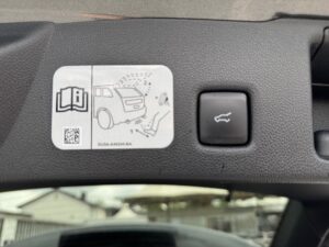 Etikett im Auto mit Anleitung für Heckklappen-Öffnungsmechanismus, Knopf daneben, grauer Hintergrund.