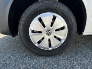 Nahaufnahme eines Autoreifens mit silberner Radkappe und VW-Logo auf Kiesboden im Sonnenlicht.