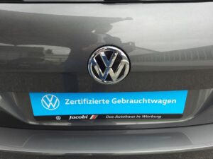 Volkswagen-Emblem auf grauem Auto mit blauem Schild "Zertifizierte Gebrauchtwagen".