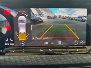 Bild eines Autodisplays mit Rückfahrkameraansicht, umgeben von parkenden Autos und Gebäuden.