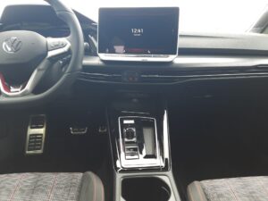 Innenansicht eines modernen Autos mit digitalem Display und Lenkrad; graue Sitze mit roten Details.