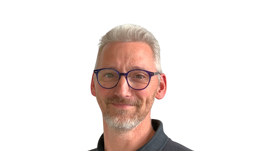 Ein lächelnder Mann mit grauem Haar trägt eine Brille und ein dunkles Hemd